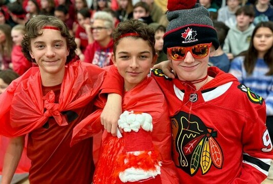 Three students all dressed in red wearing red plastic capes and red head bands. They are standing side by side with arms around each other, in the context of a school pep ralley.