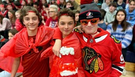 Three students all dressed in red wearing red plastic capes and red head bands. They are standing side by side with arms around each other, in the context of a school pep ralley.