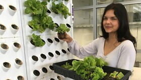 High school student standing by hydroponics machine, holding a lettuce plant in one hand and tray of plants in the other
